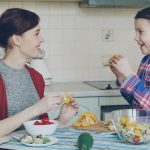 Happy family of young mother and cute cheerful daughter have fun grimacing silly with vegetables while cooking in the kitchen at home